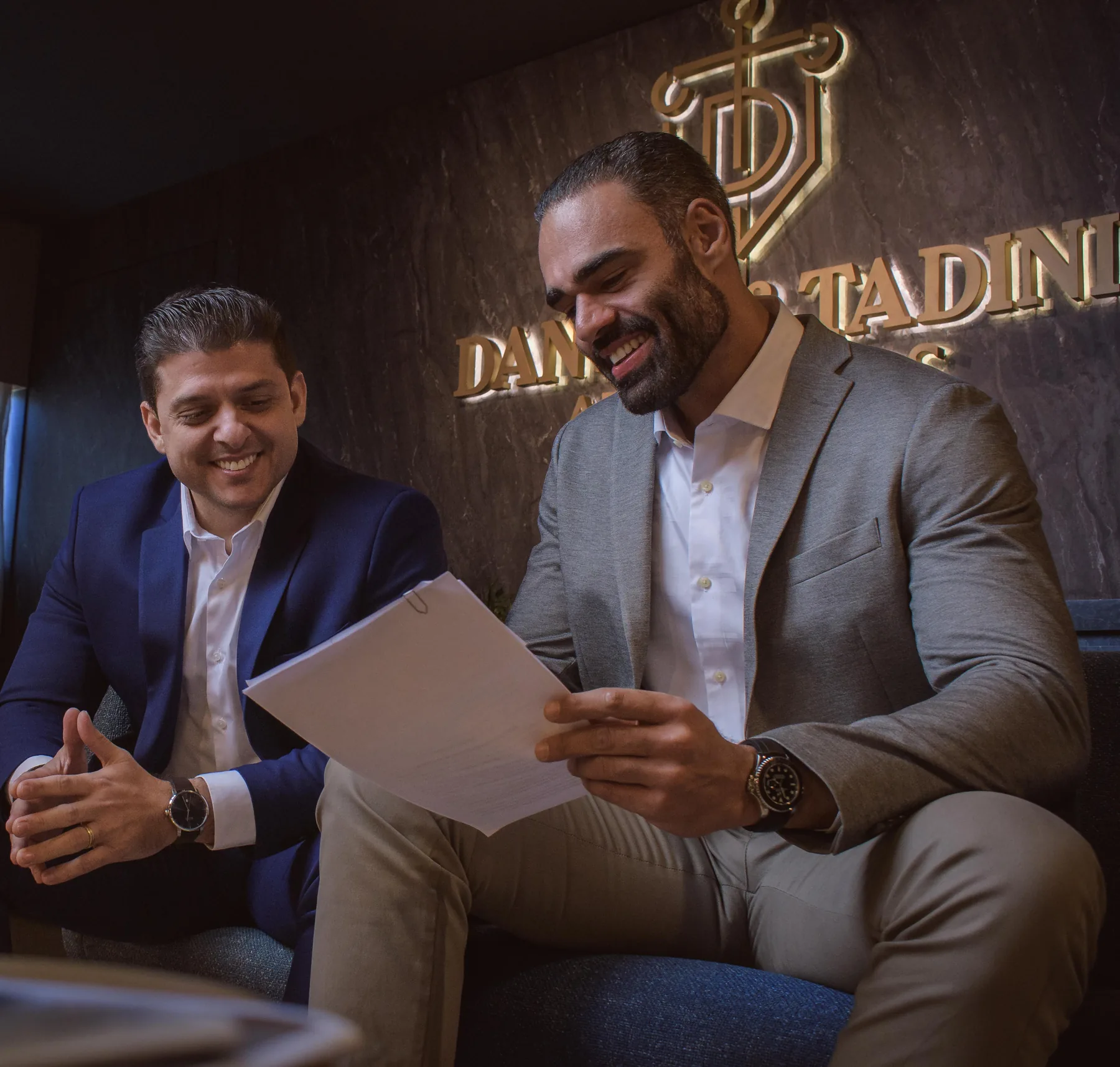 Os sócios Felipe Tadini e Gustavo Dantas sorrindo, sentados lado a lado no escritório, sob o logotipo dourado Dantas & Tadini